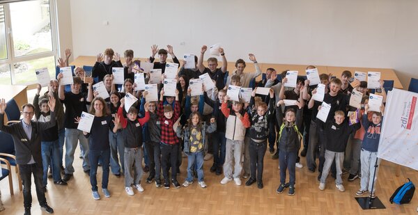 Zahlreiche Sch&uuml;lerinnen, Sch&uuml;ler und einige Lehrkr&auml;fte stehen in einem gro&szlig;en Saal, lachen, recken die H&auml;nde in die Luft und schwenken Urkunden. 