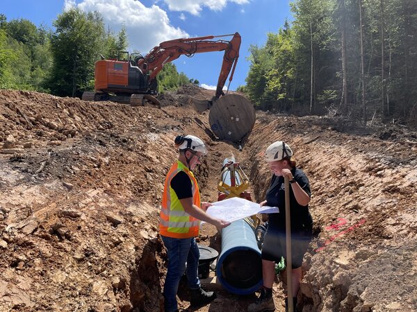 Die Personen stehen auf einer Baustelle in einer Rinne , in die bereits Wasserrohre verlegt wurden. Beide tragen Helme und betrachten einen gro&szlig;en Bauplan. Hinter ihnen steigt ein H&uuml;gel steil an, darauf ein Bagger.