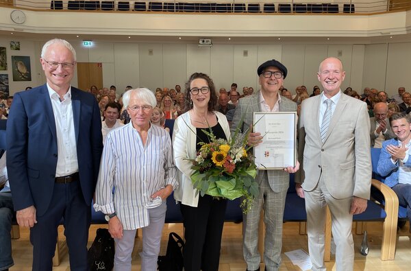 Die Personen stehen vor einem vollen, gro&szlig;en Sitzungssaal. G&auml;ste im Hintergrund applaudieren. 