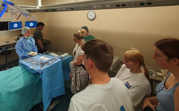 OP-Krankenpflegerin oder &Auml;rztin mit blauem Kittel und Haube steht vor medizinischen Instrumenten in einem OP. Eine Gruppe Jugendlicher schaut interessiert zu ihr.