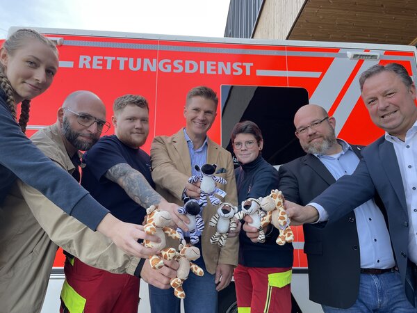 Die Personen stehen vor einem Rettungswagen im Halbkreis und zeigen Pl&uuml;schtiere in die Kamera.